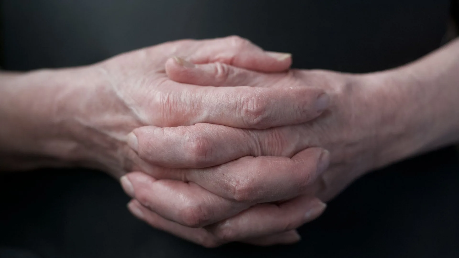 Close crop of an older person’s weathered hands folded together and resting in low light