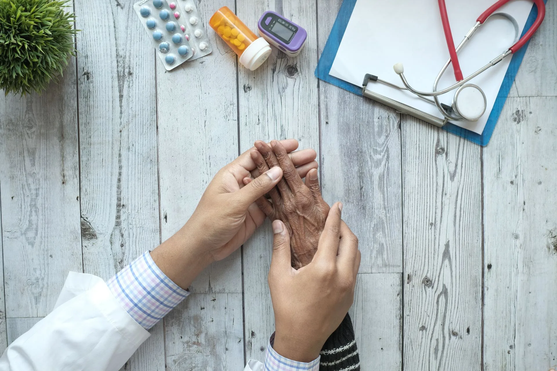 A clinician resting a hand over an older patient’s clasped hands across a desk