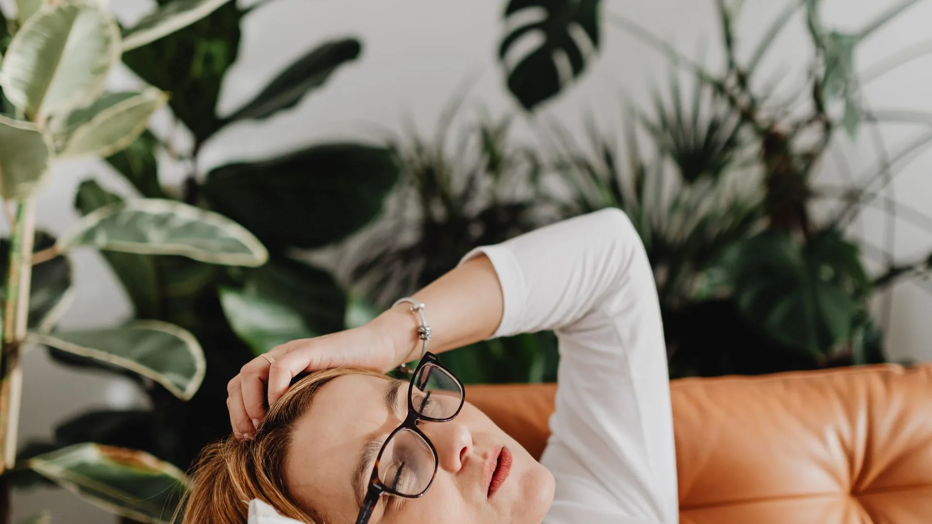 A woman lying back on a couch at home with her eyes closed, resting among houseplants in daylight