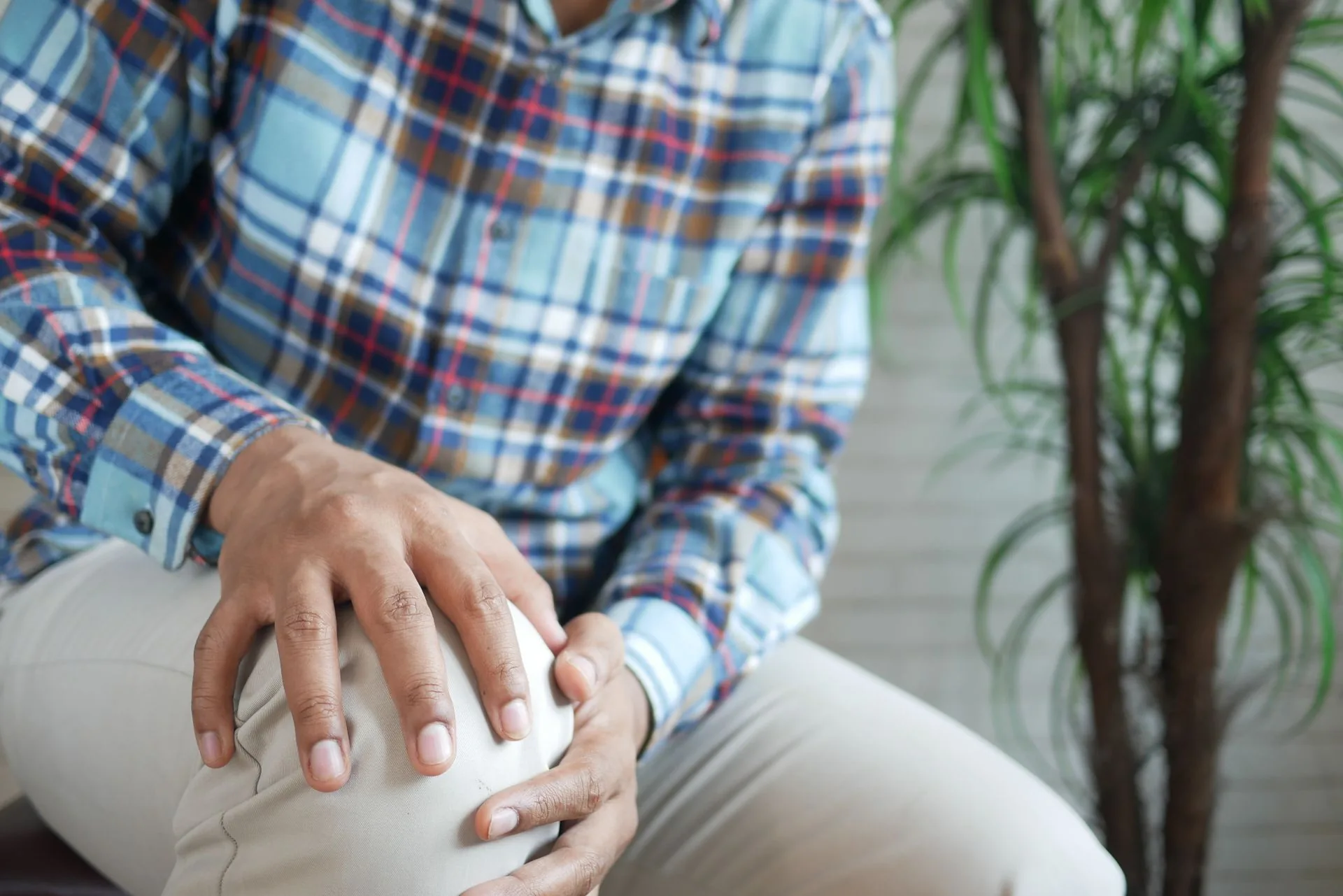 A seated man in a plaid shirt with both hands cupped over his knee