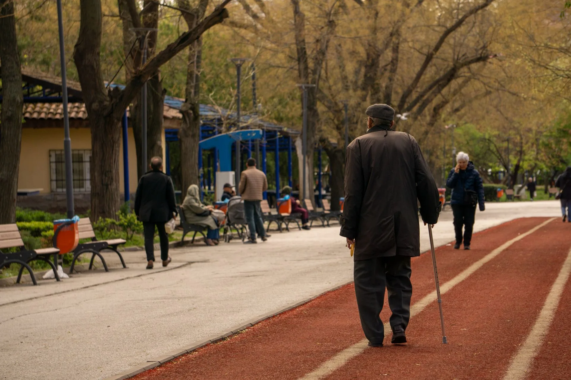 An older man with a walking cane pausing on a tree-lined park path
